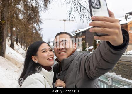 Joyful Asian couple captures a selfie amidst the winter scenery of Zermatt, surrounded by snow and festive ambiance, both looking at screen. Stock Photo
