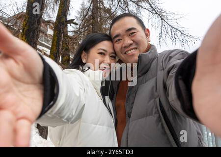 Cheerful Asian couple takes a close-up selfie in Zermatt with a snowy backdrop during Christmas. Both are looking at the camera, smiling warmly, captu Stock Photo