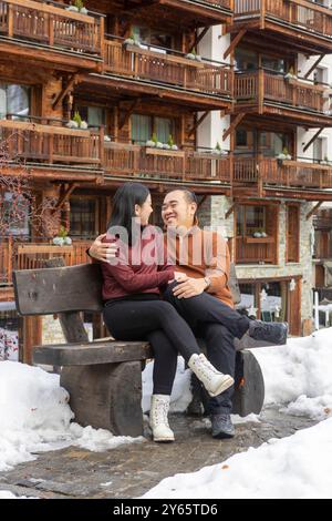 Asian couple sharing an affectionate moment on a snow-covered bench in Zermatt, looking at each other with smiles, against a charming wooden chalet ba Stock Photo
