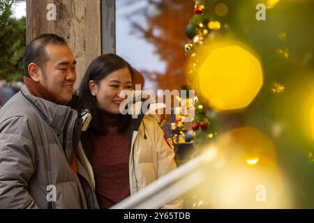 A joyous Asian couple is seen window shopping at a beautifully decorated store during Christmas time in Zermatt. Both are smiling and looking at festi Stock Photo