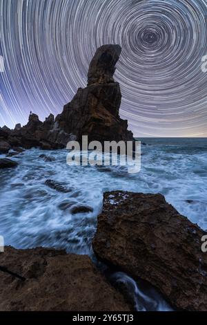 A long exposure photograph captures star trails swirling over a spectacular rock formation in Cabo de Gata, Almeria Waves crash energetically against Stock Photo