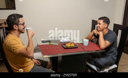 An intimate moment captured between two men on a gay date, enjoying coffee and snacks at a dining table, showcasing a loving LGBTQIA+ relationship in Stock Photo