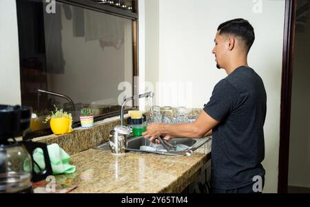 A gay man washes dishes in a well-lit kitchen with granite countertops and a window in the background He is focused on cleaning utensils, creating a r Stock Photo