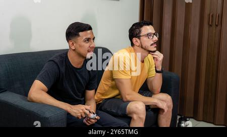 Two young men, possibly on a gay date, enjoy a relaxed gaming session together on a sofa, embodying the spirit of contemporary LGBTQIA+ relationships Stock Photo