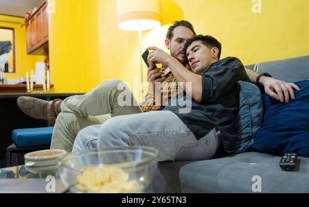 Two gay men enjoy a cozy evening on a couch, engaged in using a smartphone together, surrounded by a warmly lit living room with snacks and candles Stock Photo