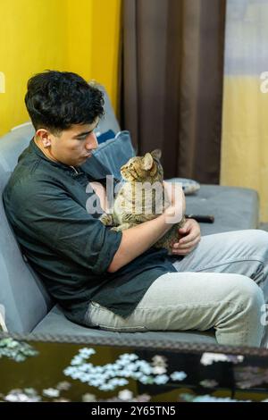 A young man sits comfortably on a grey sofa, gently holding a cat in his lap, both enjoying a peaceful moment at home with a warm yellow background Stock Photo