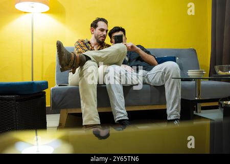 A gay couple lounges comfortably on a gray sofa, engaging with a smartphone in a warmly lit living room with a vibrant yellow wall Stock Photo
