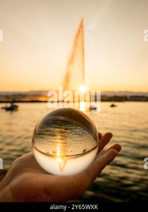 Cropped unrecognizable person's hand holding a clear crystal ball, focusing on Geneva's iconic Jet d’eau and the surrounding landscape inverted within Stock Photo