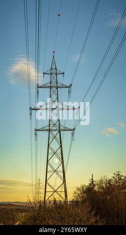 A serene sunset landscape featuring an electricity pylon rising against a softly lit sky, with subtle hints of foliage and distant hills visible in th Stock Photo