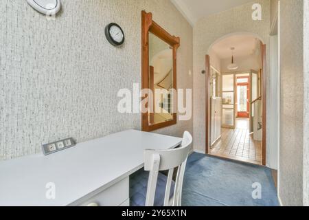 A well-lit hallway featuring a textured wall, large wooden mirror, white desk with chair, wall-mounted clock, and an open doorway. Stock Photo