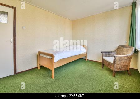 A basic bedroom featuring a single wooden bed with white bedding and a wicker armchair, with green carpeting and wallpapered walls. Stock Photo