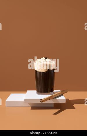 A delectable glass of coffee topped with vanilla ice cream and a cinnamon stick, presented on a white platform with a warm beige background. Stock Photo