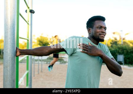 A young man stretches his arm while using outdoor exercise equipment in a sandy park area, with a backdrop of green trees and clear skies, possibly tr Stock Photo