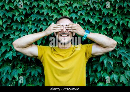 A joyful man in a yellow t-shirt covers his eyes with his hands, smiling broadly against a vibrant ivy-covered wall Stock Photo