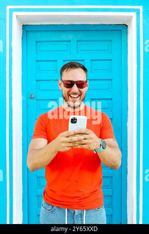 Cheerful man in orange T-shirt standing against a vivid blue door, using his smartphone with a joyful expression His modern, casual style and engaging Stock Photo