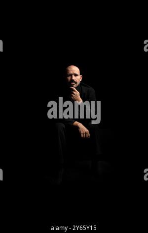 A middle-aged bald man in a black shirt, sitting alone in a dark room, seems lost in deep thought with a somber expression The obscure background enha Stock Photo