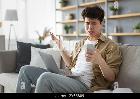 Confused asian man looking at mobile phone using laptop Stock Photo