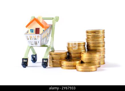 Miniature house placed in a toy shopping cart next to stacks of coins, symbolizing real estate investment, finance management, and economic growth. Stock Photo