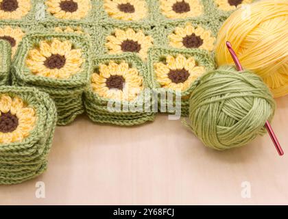 Close up on piles of finished crochet granny squares, connected squares behind with balls of yellow and green yarn to side on light wood table. Croche Stock Photo