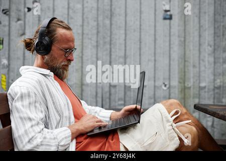 Minimal side view portrait of contemporary bearded man using laptop and wearing big headphones while working online or using internet sitting outdoors Stock Photo