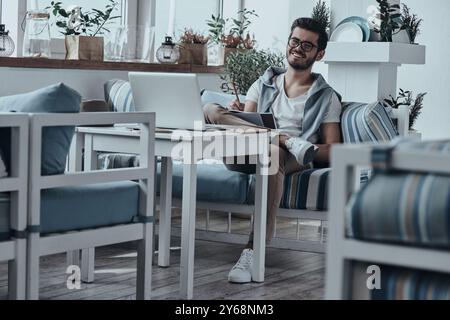 Enyoing nice day. Good looking young modern man writing something down in his notebook while sitting on the sofa in restaurant Stock Photo
