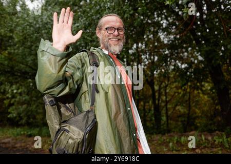 Waist up portrait of bearded adult man with backpack waving at camera and smiling while enjoying tourism in nature, copy space Stock Photo