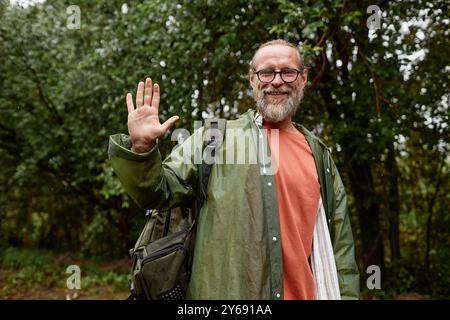 Waist up portrait of smiling senior man wearing raincoat waving at camera and enjoying tourism in nature, copy space Stock Photo