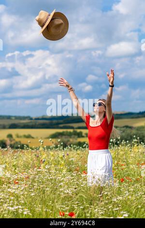 Blonde woman in sunglasses in a wildflower field throwing straw hats in the air Stock Photo