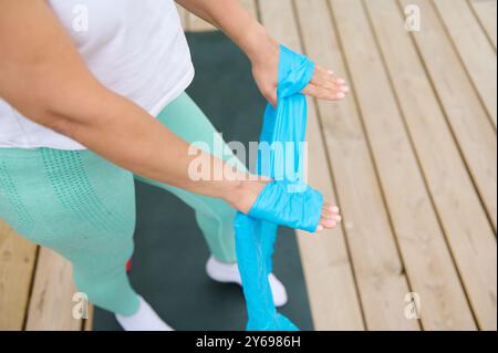 Woman using a blue resistance band for strength training exercises on a wooden floor. Focus on hands and exercise technique. Stock Photo