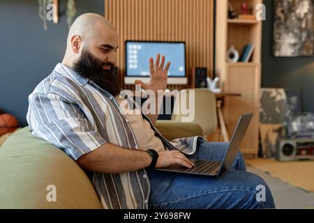 Smiling bearded man sitting on couch in modern living room, using laptop and gesturing with hand, wearing striped shirt and jeans, working from home Stock Photo
