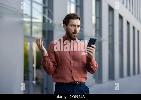 Mature male professional standing outside in city street holding phone, expression of shock and disbelief. Businessman in smart casual attire reacting to surprising news Stock Photo