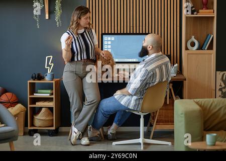 Colleagues discussing project in modern home office, with computer monitor in background while standing and sitting next to desk displaying engaged conversation Stock Photo