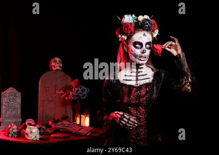 Woman with flower at Day of the Dead event with spooky decor to remember the deceased, celebrating their memory. Lady holding red rose for traditional holiday of remembrance for those who have passed Stock Photo