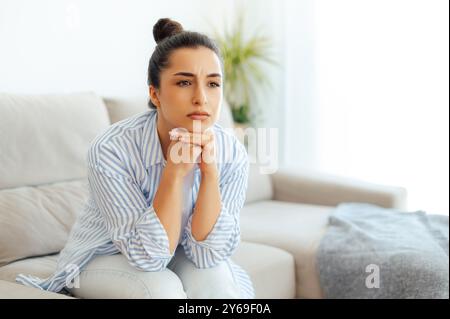 Upset arabian or indian young woman in casual clothes, sitting on a sofa in a living room, feeling sad tired and loneliness, suffering depression, worried about personal troubles or divorce, offended Stock Photo