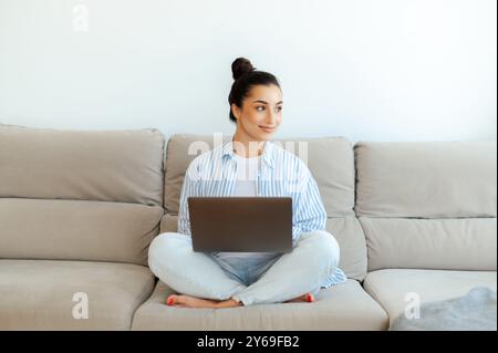 Photo of female freelancer on sofa. Motivated positive pretty indian or arabian brunette woman, sits on comfortable couch at home in living room, using her laptop for distant work on a project, smile Stock Photo