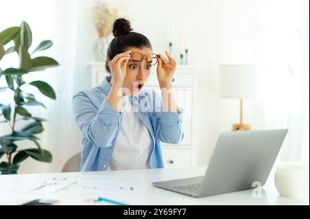 Confused shocked excited woman of arabian or indian nationality with glasses, office worker, digital manager, looks at laptop screen in surprise and puzzling lifting glasses,saw unexpected information Stock Photo
