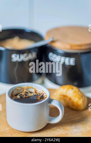 Freshly poured and steaming hot,ready to drink,coffee mug on a wooden chopping board next to a bread roll,demerara sugar and coffee containers, recent Stock Photo
