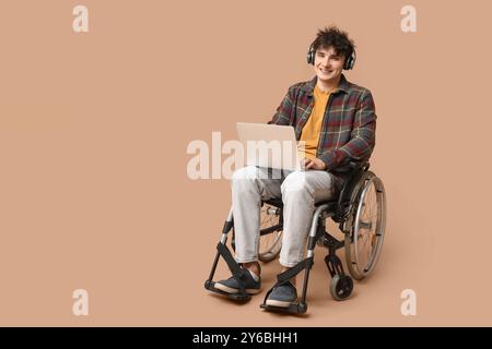 Young male programmer in headphones on wheelchair holding laptop on beige background Stock Photo