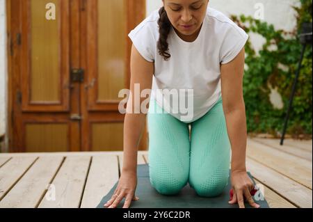 A woman kneels on a yoga mat outdoors, practicing yoga for mindfulness and relaxation. She wears a white shirt and mint green leggings on a wooden dec Stock Photo