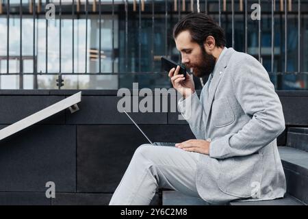 Bearded man in gray suit and sitting on steps, engaging in conversation on smartphone and using open laptop. Modern building in background creating professional atmosphere Stock Photo