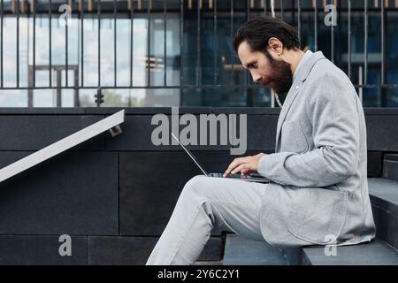 Man with beard wearing gray suit, working on laptop while sitting on outdoor steps. Reflected building glass and modern architecture in background is visible Stock Photo