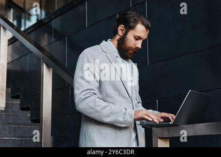 Man with thick beard standing on the stairs while using a laptop, focused on screen, dressed in formal gray suit, with one hand resting on the railing Stock Photo