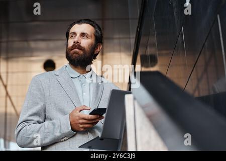 Bearded man in gray suit holding smartphone while standing in sleek, contemporary office setting with glass paneling. He has thoughtful expression on his face Stock Photo