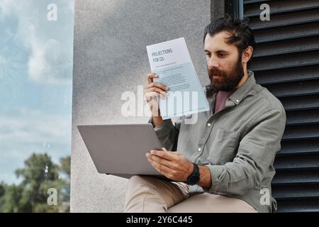 Bearded man sitting outside, holding a paper document, and using a laptop for work, surrounded by greenery and modern architecture Stock Photo