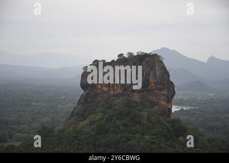 Sigiriya Rock Fortress in Sri Lanka surrounded by lush greenery and distant mountains under a cloudy sky. Stock Photo