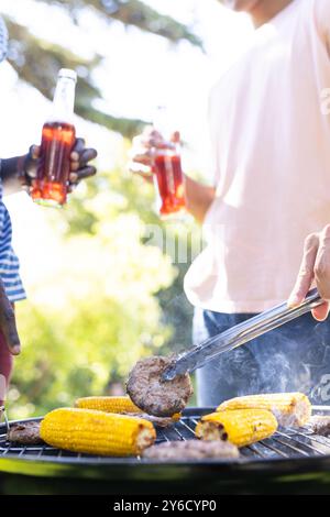 Grilling burgers and corn, two diverse male friends holding drinks and enjoying barbecue Stock Photo