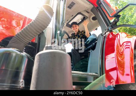 Female farmer sitting in tractor cabin smiling and waving. Woman driving agricultural or construction tractor. Stock Photo