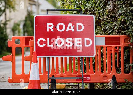 Red Road Closed Ahead sign with white lettering, with an orange plastic barrier and an orange and white road cone on a rural country road Stock Photo