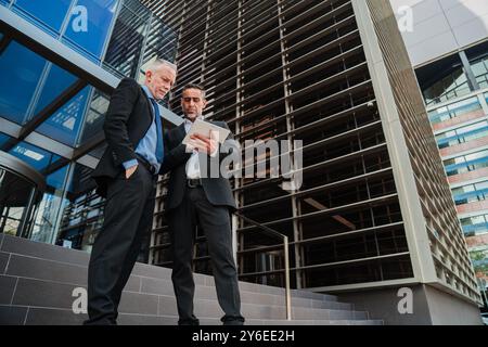 Two businessmen standing outside a modern office building, using a tablet for discussing work, collaborating on a business project, and reviewing important documents in a professional setting. High quality photo Stock Photo