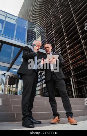 Vertical Two businessmen standing outside a modern office building, using a tablet for discussing work, collaborating on a business project, and reviewing important documents in a professional setting. High quality photo Stock Photo